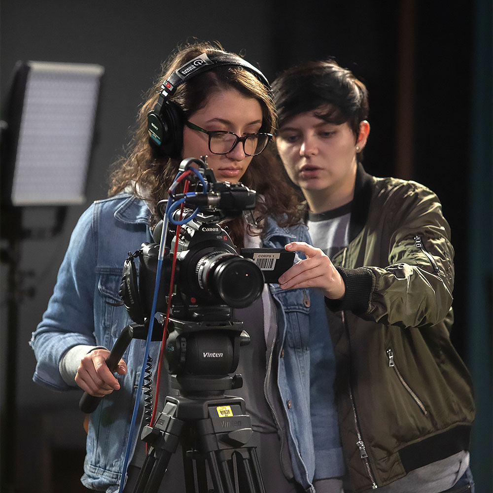 Two video production students stand behind a professional video camera.