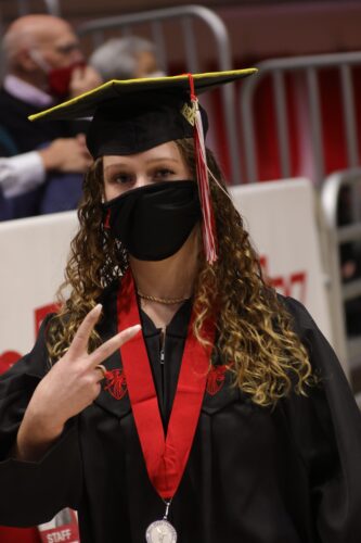 A woman with a cap n' gown holding up two finers in a peace sign position with a mask on.